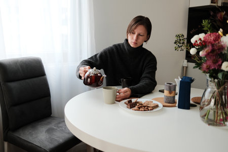 A woman pours tea into a mug while sitting at a round table with snacks beside her. The scene captures a peaceful break and a warm daily ritualの写真素材