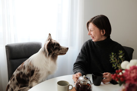 A woman laughs while holding a teapot as her dog curiously watches, capturing a warm and playful domestic interaction. The scene shows companionship and relaxed morning moodの写真素材