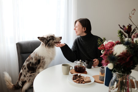 A woman sits at a table smiling warmly at her dog as it looks up at her. Concept of cozy domestic morning routineの写真素材