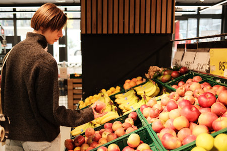 A woman holds an apple while standing at the fruit display in a grocery store. The concept shows everyday shopping and healthy food selectionの写真素材