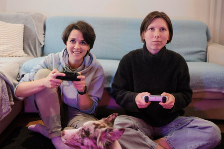 Two women sit on the floor playing a video game while their dog watches closely. The moment captures friendly competition and a cozy, relaxed home moodの写真素材