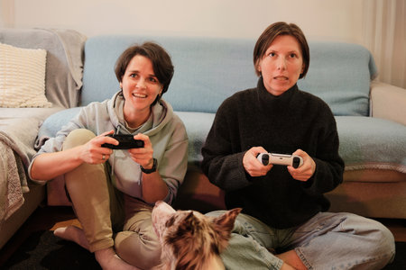Two women sit on the floor playing a video game while their dog watches closely. The moment captures friendly competition and a cozy, relaxed home moodの写真素材