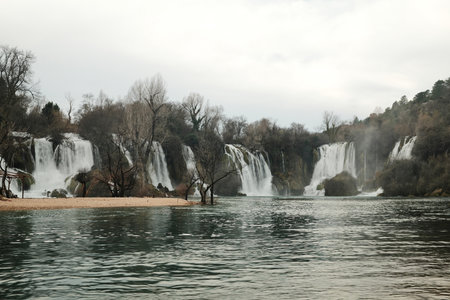 Wide cascading Kravica Waterfall surrounded by forest and turquoise river water in Bosnia and Herzegovina. Natural landscape with flowing streams, mist and calm atmosphere in early springの写真素材