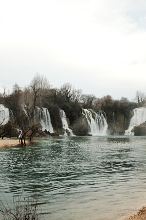 Wide cascading Kravica Waterfall surrounded by forest and turquoise river water in Bosnia and Herzegovina. Natural landscape with flowing streams, mist and calm atmosphere in early springの写真素材