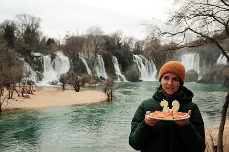 Smiling woman holding a cake with candles in front of Waterfall. Travel celebration concept with nature landscape, winter atmosphere, and joyful moodの写真素材