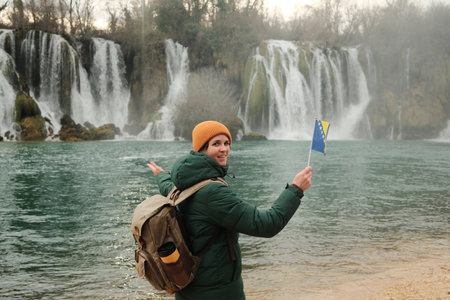 Smiling female traveler turning back and presenting Kravica Waterfall while holding flag. Travel storytelling concept with nature scenery and winter outdoor moodの写真素材