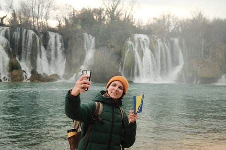 Woman taking selfie or making video while holding Bosnia and Herzegovina flag in front of Kravica Waterfall. Modern travel lifestyle with smartphone, nature scenery, winter mood, and personal memoryの写真素材