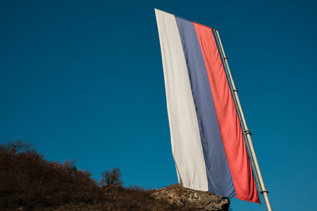 Serbian national flag waves on a tall pole above rocky hillside near Visegrad, Bosnia and Herzegovina. Clear blue sky and strong vertical composition create a calm patriotic sceneの写真素材