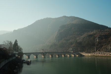 Stone bridge crosses the Drina River with forested hills rising above Visegrad, Bosnia and Herzegovina. Soft morning light creates a calm and atmospheric mountain landscapeの写真素材