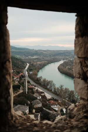Winding Neretva river and rural road seen from a narrow stone opening inside Pocitelj Fortress, flowing through a quiet valley with villages and distant mountainsの写真素材