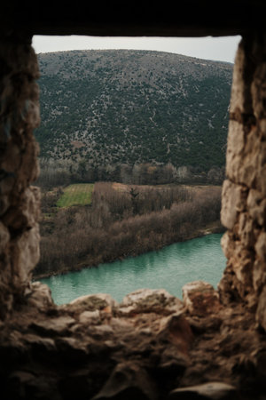 Turquoise Neretva river flowing beneath forested hills and open fields, viewed through rough stone walls of Pocitelj Fortress in a calm natural landscapeの写真素材