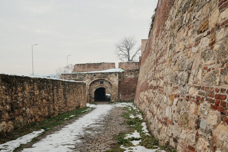 Cobblestone road between high stone and brick fortress walls leading toward an arched gate, with traces of snow, emphasizing historic urban defense architectureの写真素材
