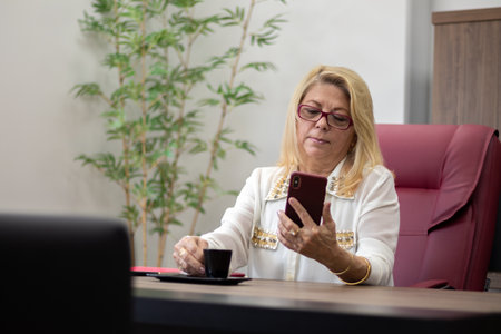 executive lady sitting at her desk looking at her cell phone, 60 year old woman, woman in charge, blonde lady in her officeの写真素材