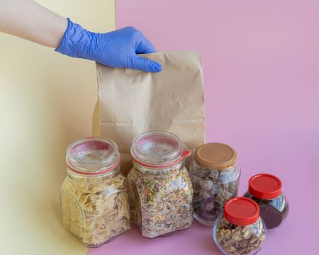 Volunteer hand in protective glove holds paper bag with different food on a pink and yellow...