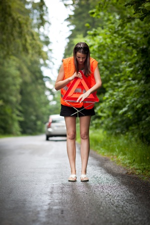 Young female driver wearing a high visibility vest/safety vest, putting in place the warning triangle after her car has broken downの写真素材