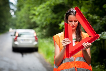 Young female driver wearing a high visibility vest/safety vest, putting in place the warning triangle after her car has broken downの写真素材