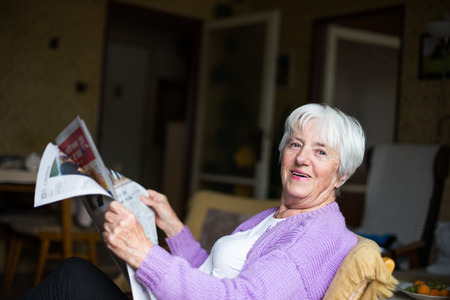 Senior woman reading morning newspaper, sitting in her favorite chair in her living room, looking happyの写真素材