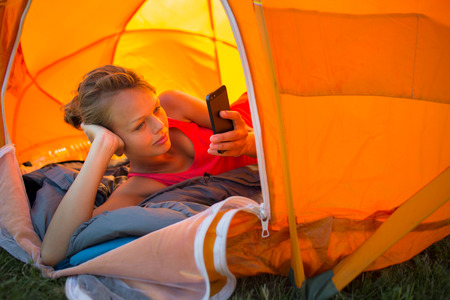 Pretty, young woman camping outdoors, lying in the tent in the morning, using her smart phoneの写真素材