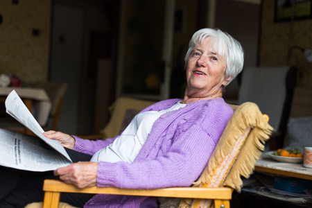 Senior woman reading morning newspaper, sitting in her favorite chair in her living room, looking happyの写真素材