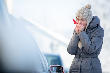 Young woman cleaning her car from snow and frost on a winter morning, she is freezing and needs to...