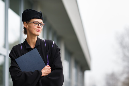 Pretty, young woman celebrating joyfully her graduation - cheking her diploma, happy/impressed with the title she received (color toned image; shallow DOF)の写真素材