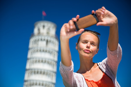 Gorgeous young woman taking a selfie with her smart phone in front of the Leaning Tower of Pisa, Tuscany, Italy (shallow DOF)の写真素材