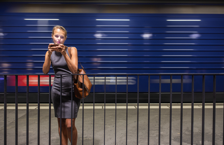 Pretty, young female commuter waiting for her daily train in a modern trainstation, using her cellphone while waiting (color toned image)の写真素材