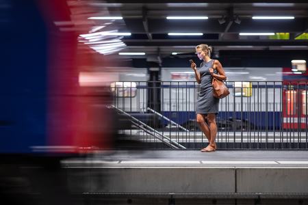 Pretty, young female commuter waiting for her daily train in a modern trainstation, using her cellphone while waiting (color toned image)の写真素材