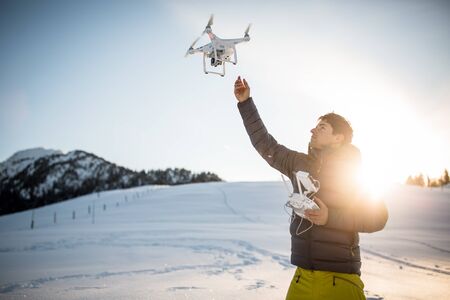 Young man controlling his drone in snowy outdoors. Drone operator holding a transmitter and landing with a drone.の写真素材