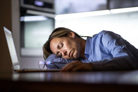 Middle-aged woman working late in the day on a laptop computer at home, running a business from home/working remotely for a corporationの写真素材