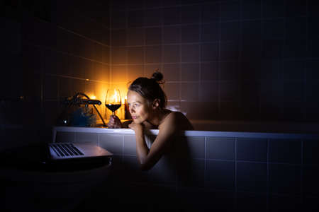 Young woman taking a bath, drinking red wine, enjoying a movie, relaxing after busy work dayの写真素材