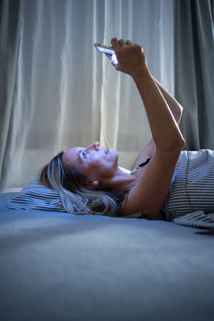 Pretty, middle-aged woman using a tablet computer in bed at home before sleep, exposing herself to bluelightの写真素材