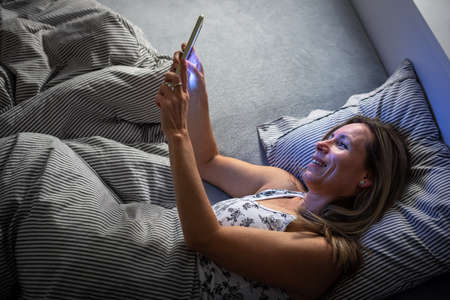 Pretty, middle-aged woman using her tablet computer before sleep in bed in the eveningの写真素材