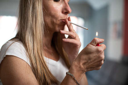 Mid-aged woman lighting a cigarette at home, getting her nicotine daily dose, unable to resist to her unhealthy habitの写真素材
