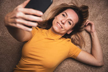 Happy mid-aged woman taking selfie with her smart phone at homeの写真素材