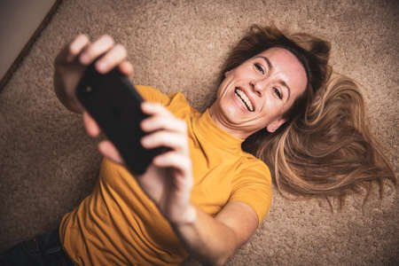 Happy mid-aged woman taking selfie with her smart phone at homeの写真素材