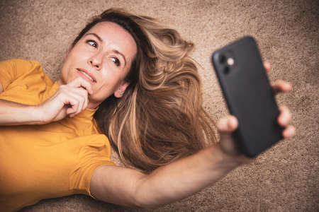 Happy mid-aged woman taking selfie with her smart phone at homeの写真素材