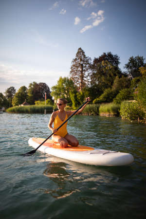 SUP Stand up paddle board concept - Pretty, young woman paddle boarding on a lovely lake in warm late afternoon lightの写真素材