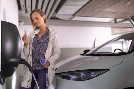 Young woman charging an electric vehicle in an underground garage equiped with e-car charger. Car sharing concept.の写真素材