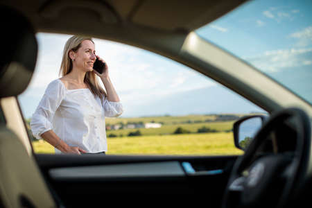 Pretty midle aged woman at the steering wheel of her car, having a break on a long trip, making a phone callの写真素材