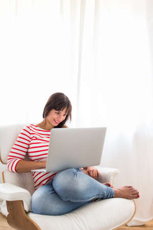 Successful young business woman sitting in a designer chair, working from home. Portrait of happy entrepreneur working on a laptop computer. Doing business remotely from the comfort of her home.の写真素材