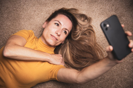 Happy mid-aged woman taking selfie with her smart phone at homeの写真素材