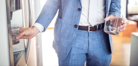 Young man in a suit pouring cold water and ice cubes from dispenser of a home fridgeの写真素材