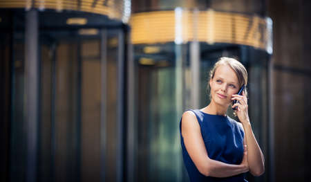 Gorgeous young businesswoman calling on her phone in front of her company's building (shallow DOF; color toned image)の写真素材