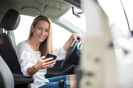 Pretty middle aged woman having at the wheel of her car, using a smartphoneの写真素材