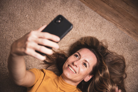 Happy mid-aged woman taking selfie with her smart phone at homeの写真素材