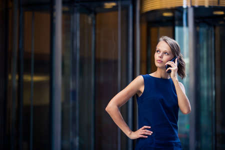 Gorgeous young businesswoman calling on her phone in front of her company's building (shallow DOF; color toned image)の写真素材