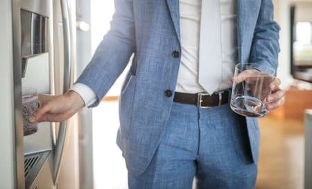 Young man in a suit pouring cold water and ice cubes from dispenser of a home fridgeの写真素材