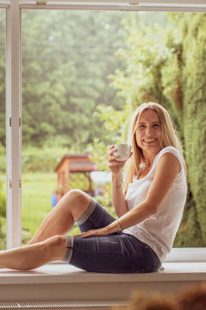 Mid-aged woman enjoying a cup of coffee in a window of her appartmentの写真素材