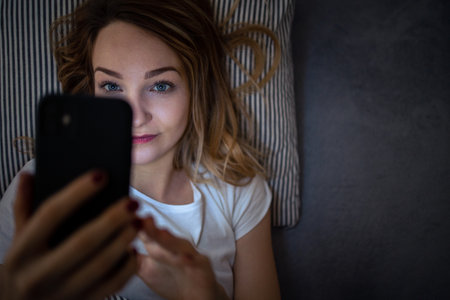 Young woman in bed holding a phone, tired and exhausted, blue light straining her eyes, messing up her circadian rhytmの写真素材